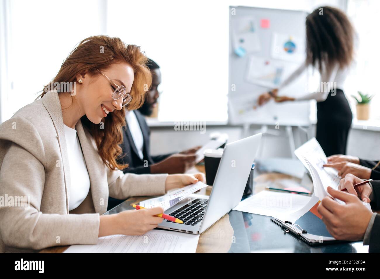 Signora di successo elegante in occhiali è seduta in ufficio a riunione di affari, l'apprendimento di grafici finanziari. Bella giovane dipendente adulta femmina facendo documenti, sorridendo Foto Stock