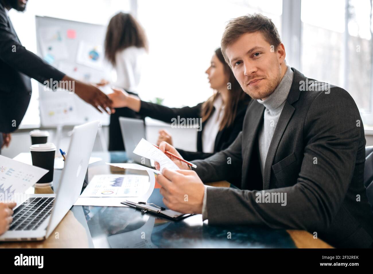 Giovane uomo d'affari caucasico in riunione di briefing, seduto al banco di lavoro, sta imparando grafici finanziari. Bell'impiegato di sesso maschile che guarda la macchina fotografica, sorridendo, facendo documenti Foto Stock