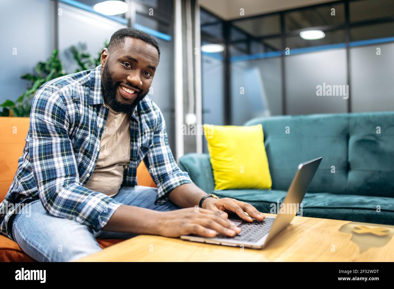 Allegro ed elegante freelancer seduto sul divano, utilizzando un computer portatile. Buon programmatore afro-americano guardando la telecamera, sorridendo, lavorando su un nuovo progetto o app Foto Stock