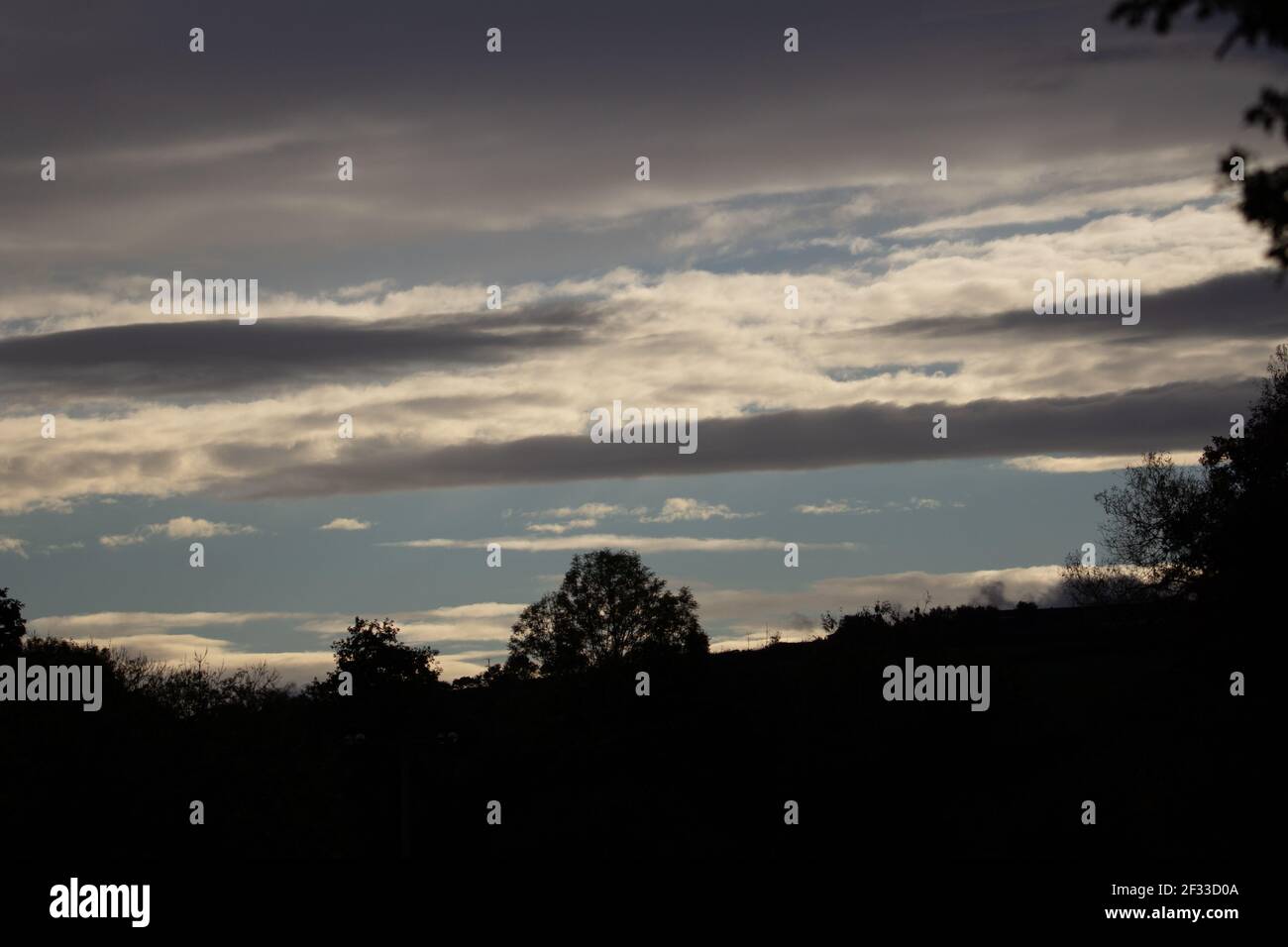 silhouette di alberi e una collina contro le nuvole e. cielo azzurro chiaro in serata Foto Stock