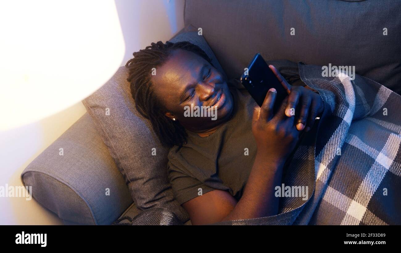 Felice giovane uomo nero sorridente mentre si usa lo smartphone. Connessione durante la quarantena. Foto di alta qualità Foto Stock