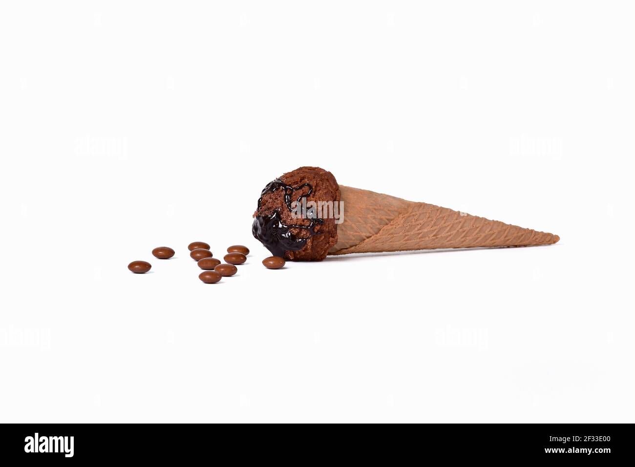 Un cono di gelato al cioccolato con sciroppo sdraiato vicino ad alcuni bottoni di cioccolato su uno sfondo chiaro. Messa a fuoco selettiva e primo piano. Concetto di gelato. Foto Stock
