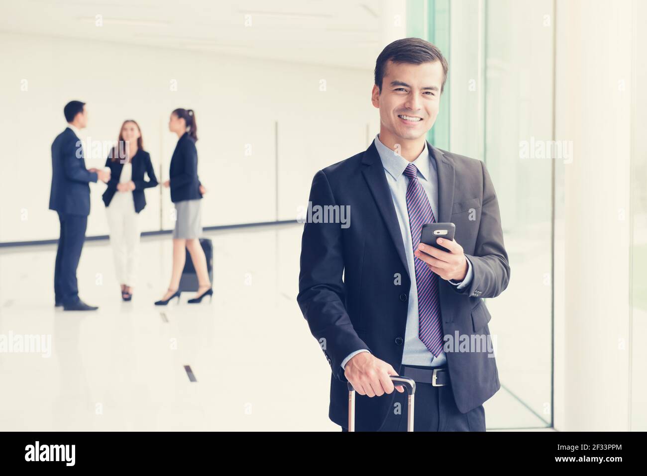 Uomo d'affari sorridente che usa lo smartphone mentre tiene la maniglia del bagaglio in un'altra mano al corridoio dell'edificio (terminal dell'aeroporto), tono vintage Foto Stock