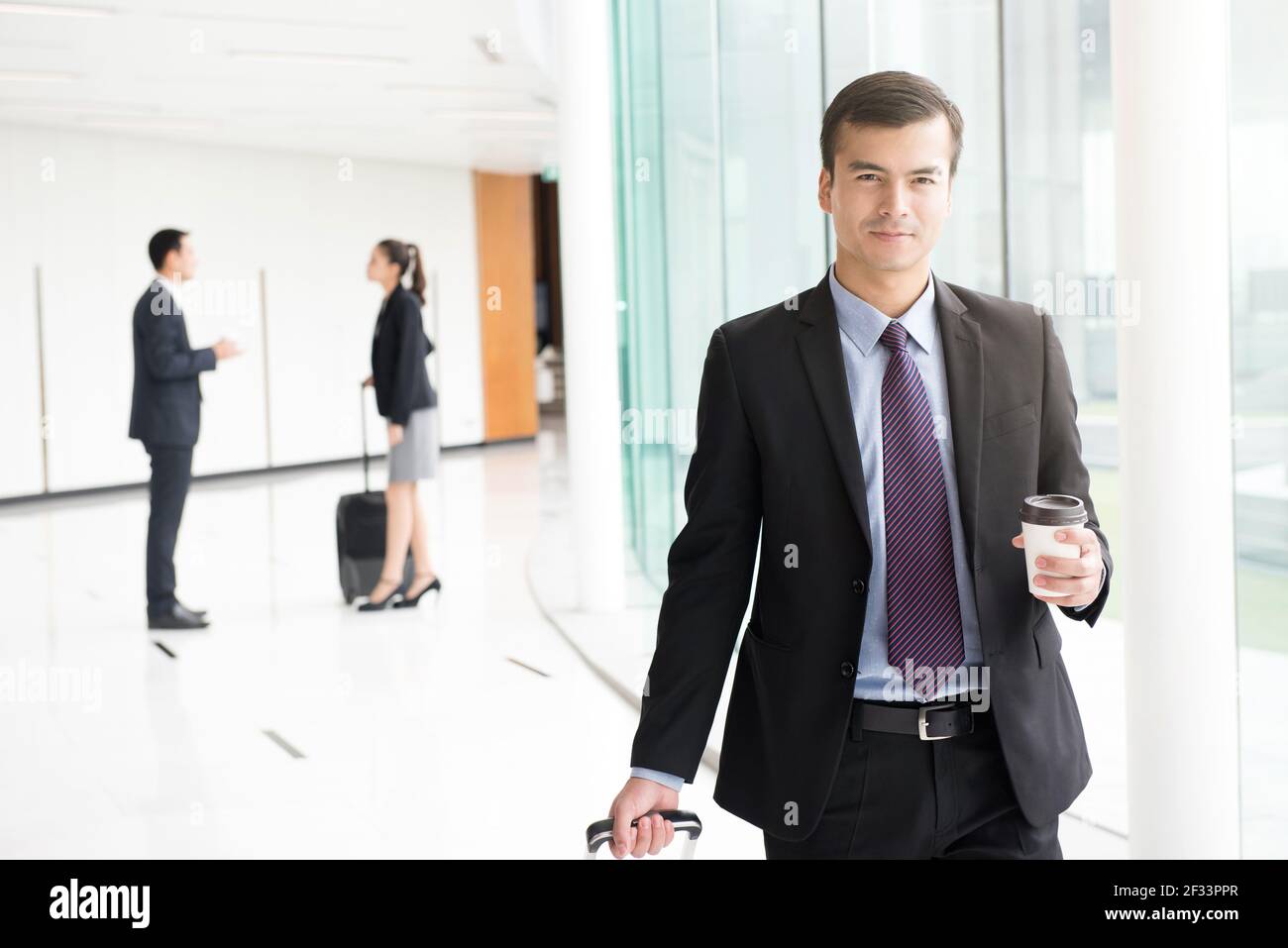 Uomo d'affari che tira il bagaglio e porta la tazza di caffè mentre si cammina corridoio dell'edificio (terminal dell'aeroporto) Foto Stock