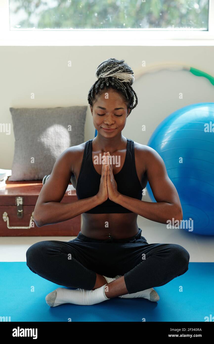 Sorridente calma giovane donna nera seduta sul tappetino yoga dentro lotus posizione e meditare con le mani nel gesto di namaste Foto Stock