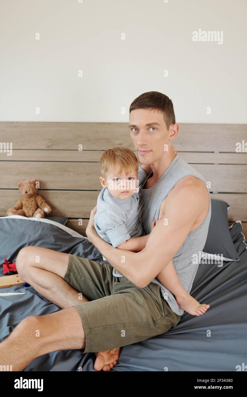 Ritratto del giovane padre che abbracciava il bambino mentre sedevano sul letto in camera da letto Foto Stock