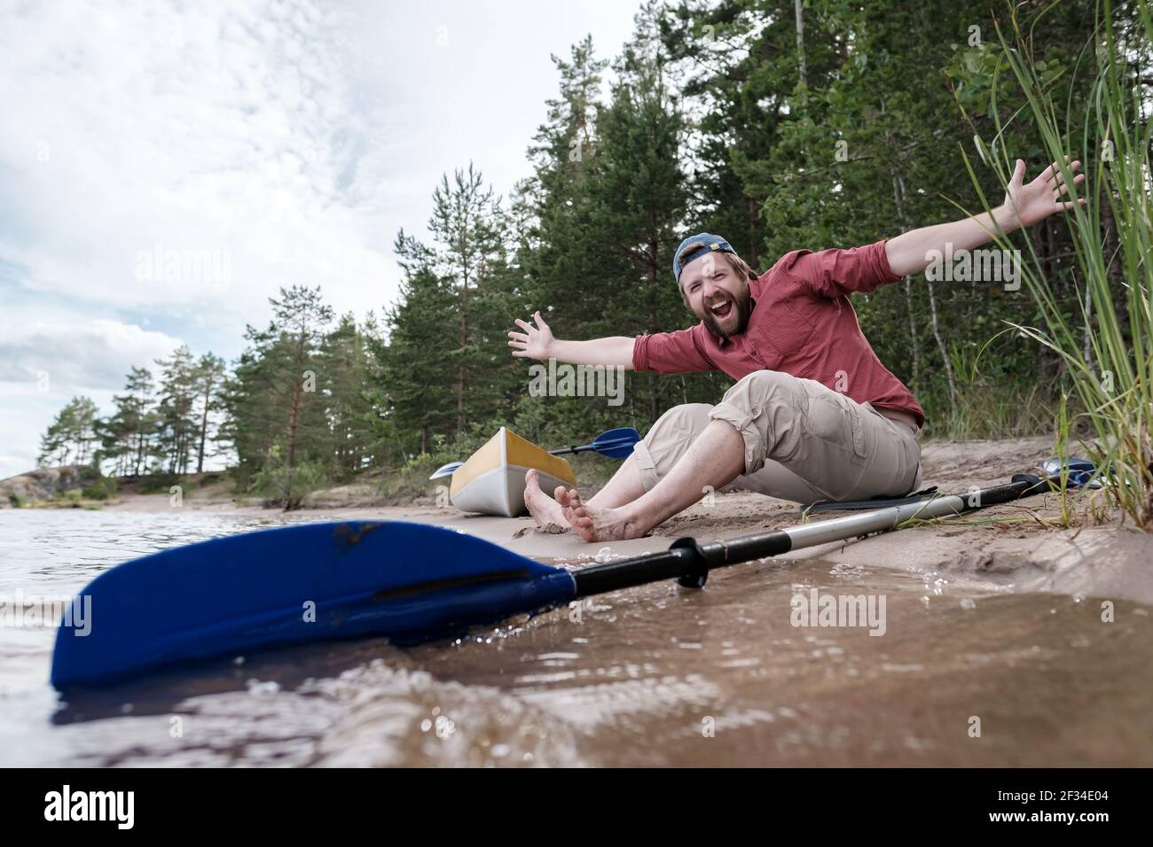 L'uomo allegro e sorridente riposa sulla riva del lago dopo un viaggio in kayak, stese le braccia ai lati e gode della natura. Foto Stock