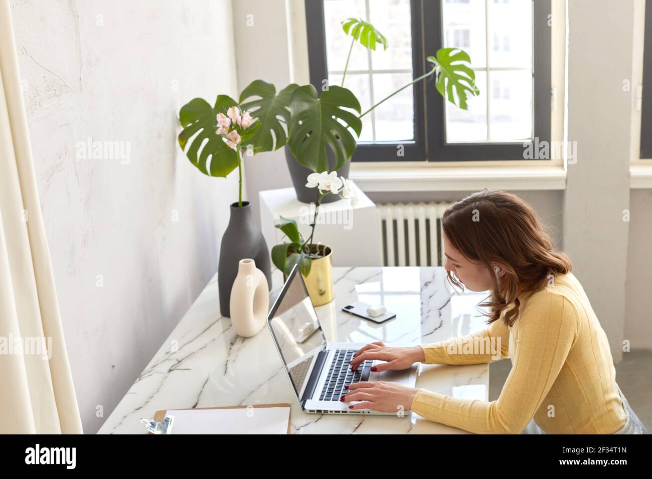 Donna d'affari in dolcevita giallo con computer portatile e seduto dal tavolo in ufficio domestico Foto Stock