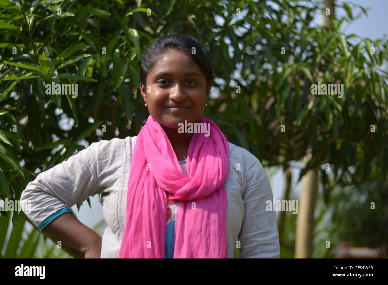 Primo piano di indios Bengalese adolescente ragazza che indossa bianco cotone salwar con dhupatta rosa in piedi di fronte a un albero di mango, messa a fuoco selettiva Foto Stock