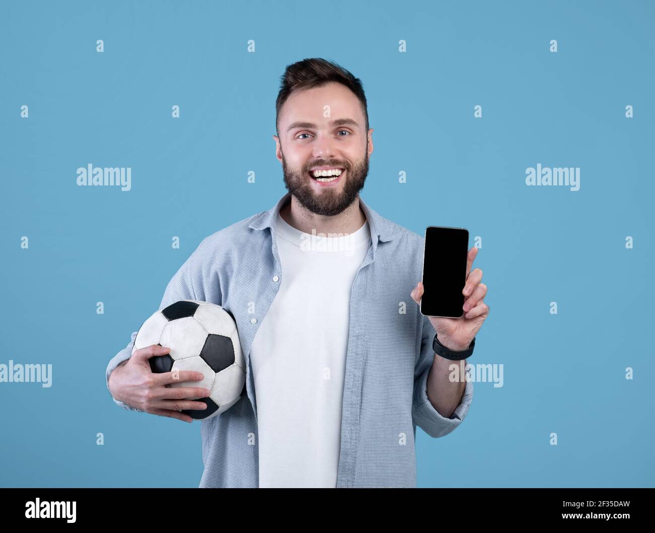 Scommettere su sport online. Ragazzo giovane che tiene la palla di calcio e il cellulare con schermo vuoto su sfondo blu, mockup Foto Stock