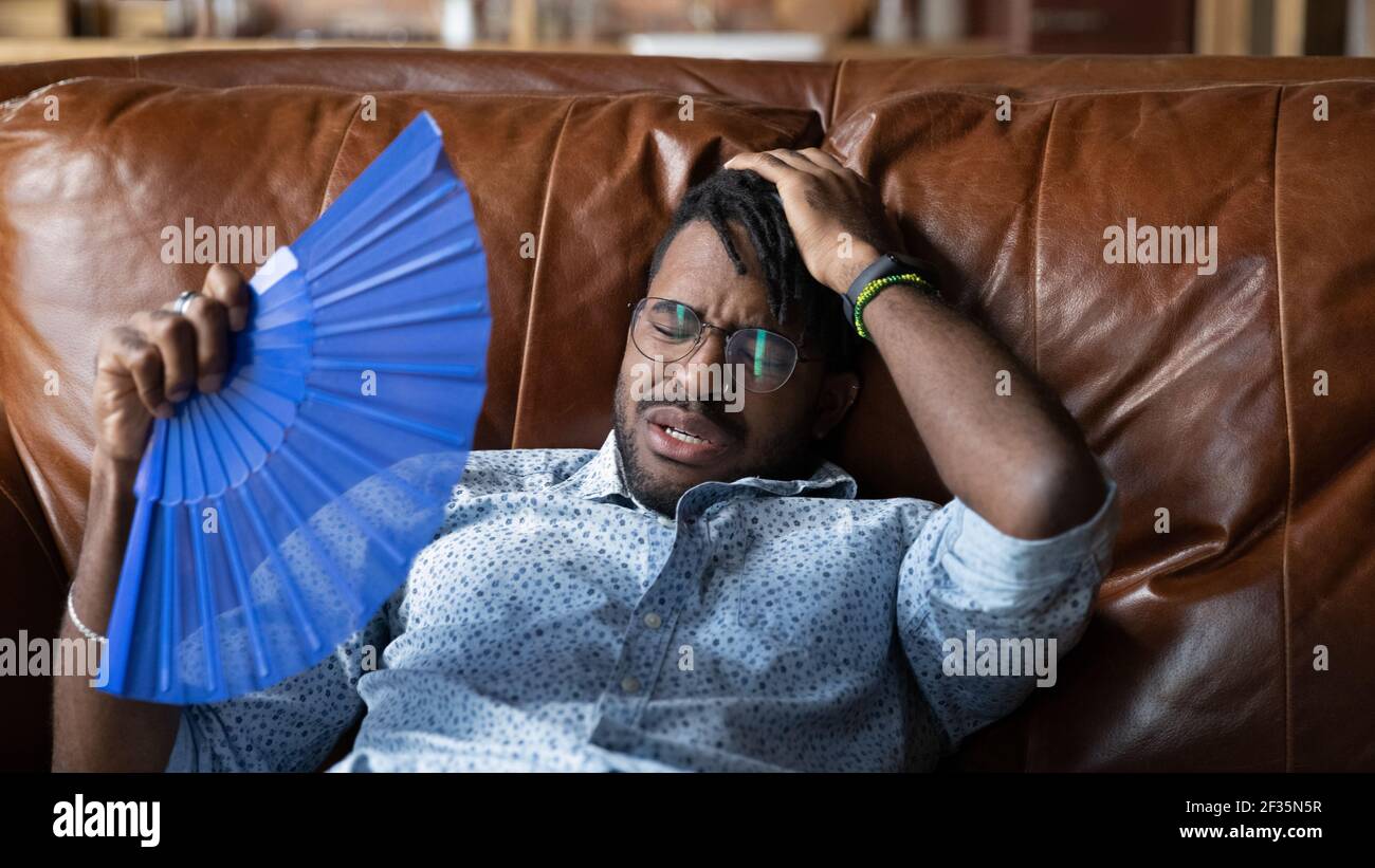 Scaldato afro american man reclinarsi sul divano utilizzare la ventola manuale Foto Stock