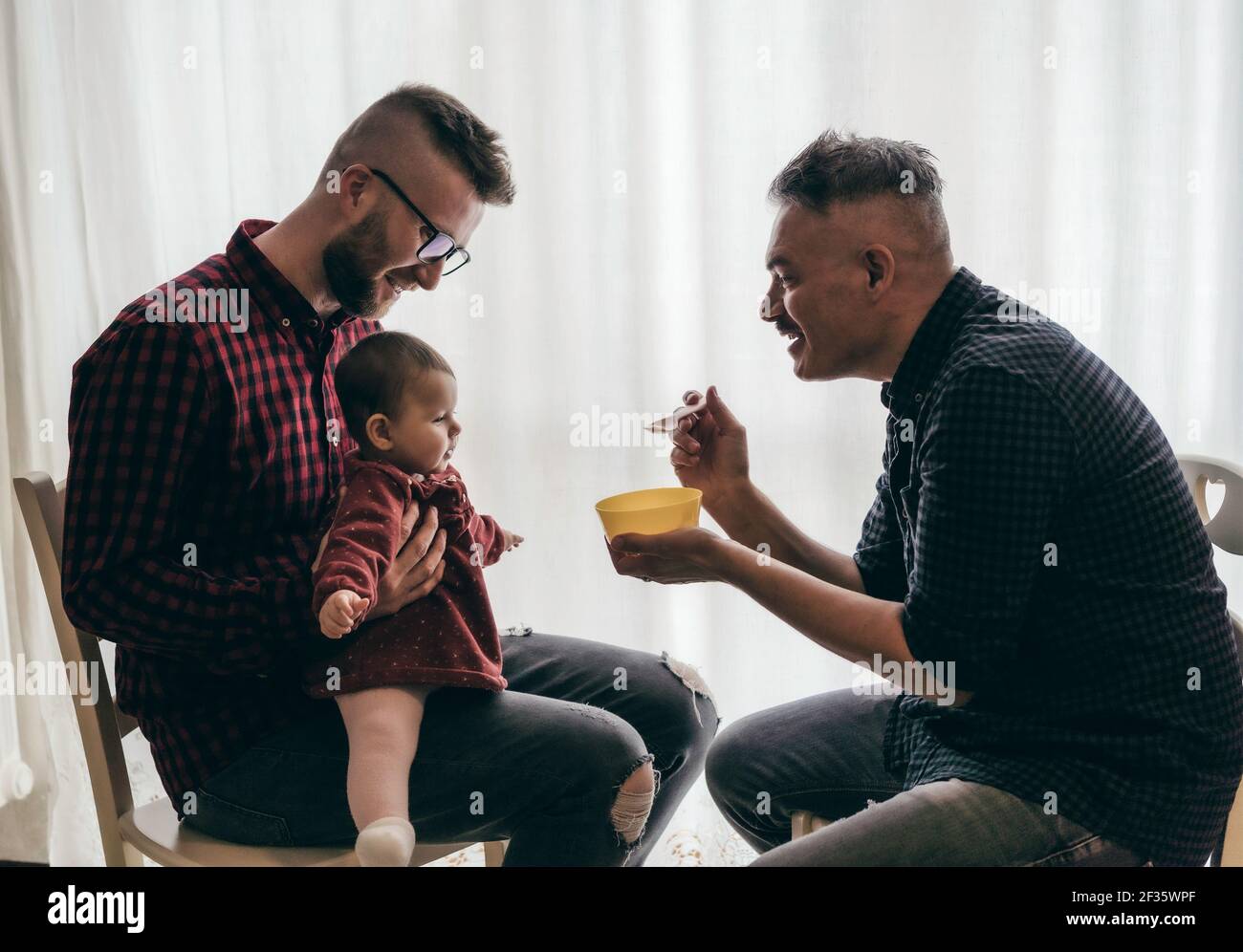 Coppia gay maschile con bambina adottata a casa - Due bei dads alimentano la ragazza del bambino sulla cucina - Babysitter maschile - la famiglia LGBT a casa - Diversity Foto Stock