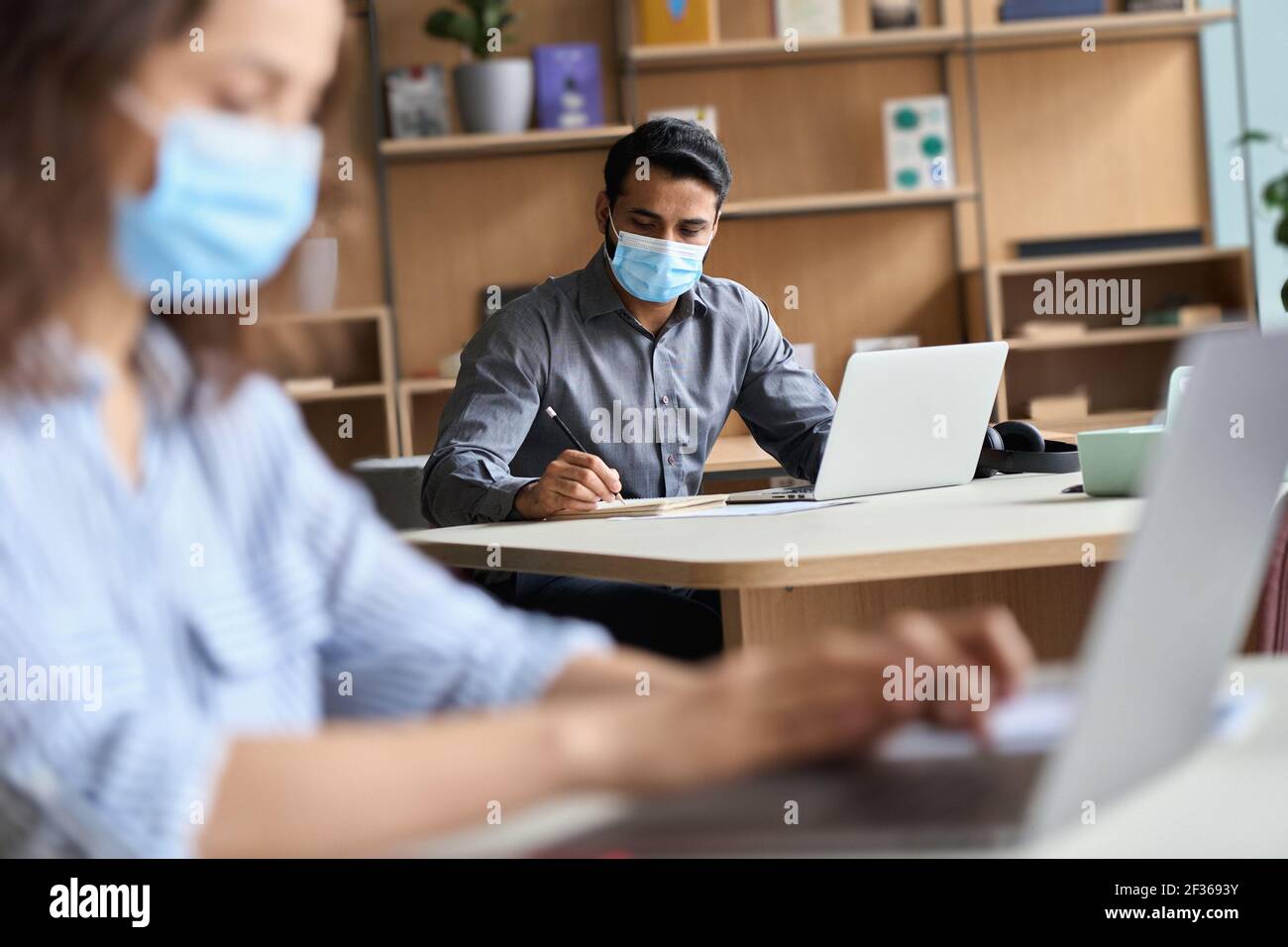 Uomo d'affari indiano che indossa la maschera facciale che lavora su un computer portatile in ufficio coworking. Foto Stock