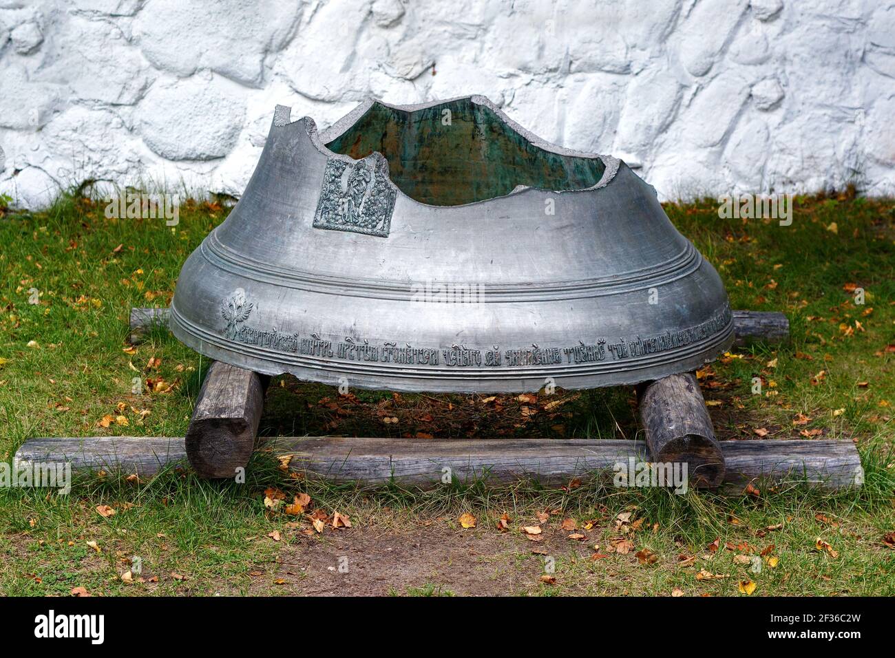 Vecchia campana della chiesa spezzata, in piedi su un basamento di tronchi di legno contro lo sfondo di una parete bianca. Foto Stock