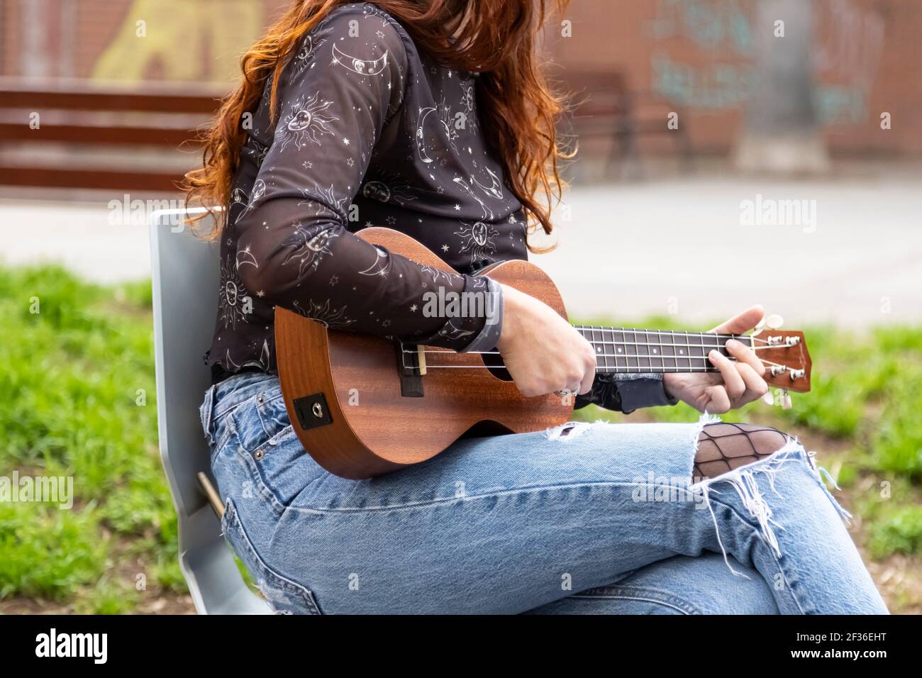 Caucasica ragazza che suona la chitarra all'aperto seduta su una sedia.  Indossa jeans e una camicia casual e porta il suo telefono cellulare nella  tasca posteriore del tr Foto stock - Alamy