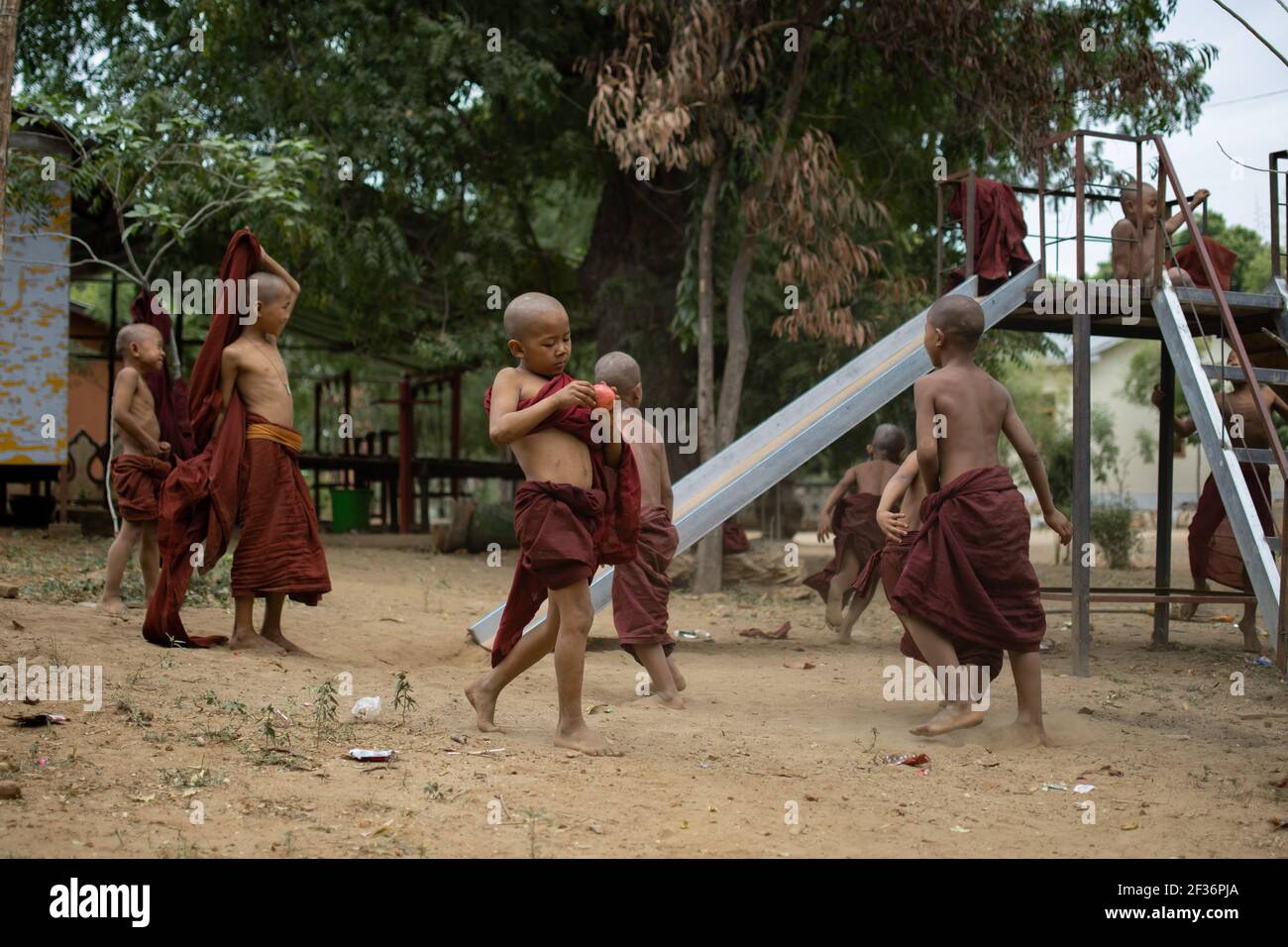 BAGAN, NYAUNG-U, MYANMAR - 2 GENNAIO 2020: Un giovane monaco mangia e mela, mentre gli altri bambini giocano e si divertono in un parco giochi locale con uno scivolo Foto Stock