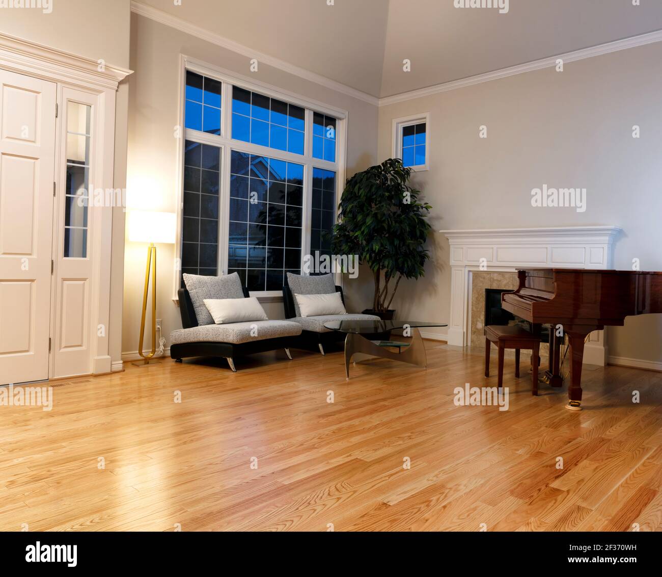 Moderno soggiorno con pavimenti in legno di quercia, piano, camino e grandi finestre durante la sera Foto Stock
