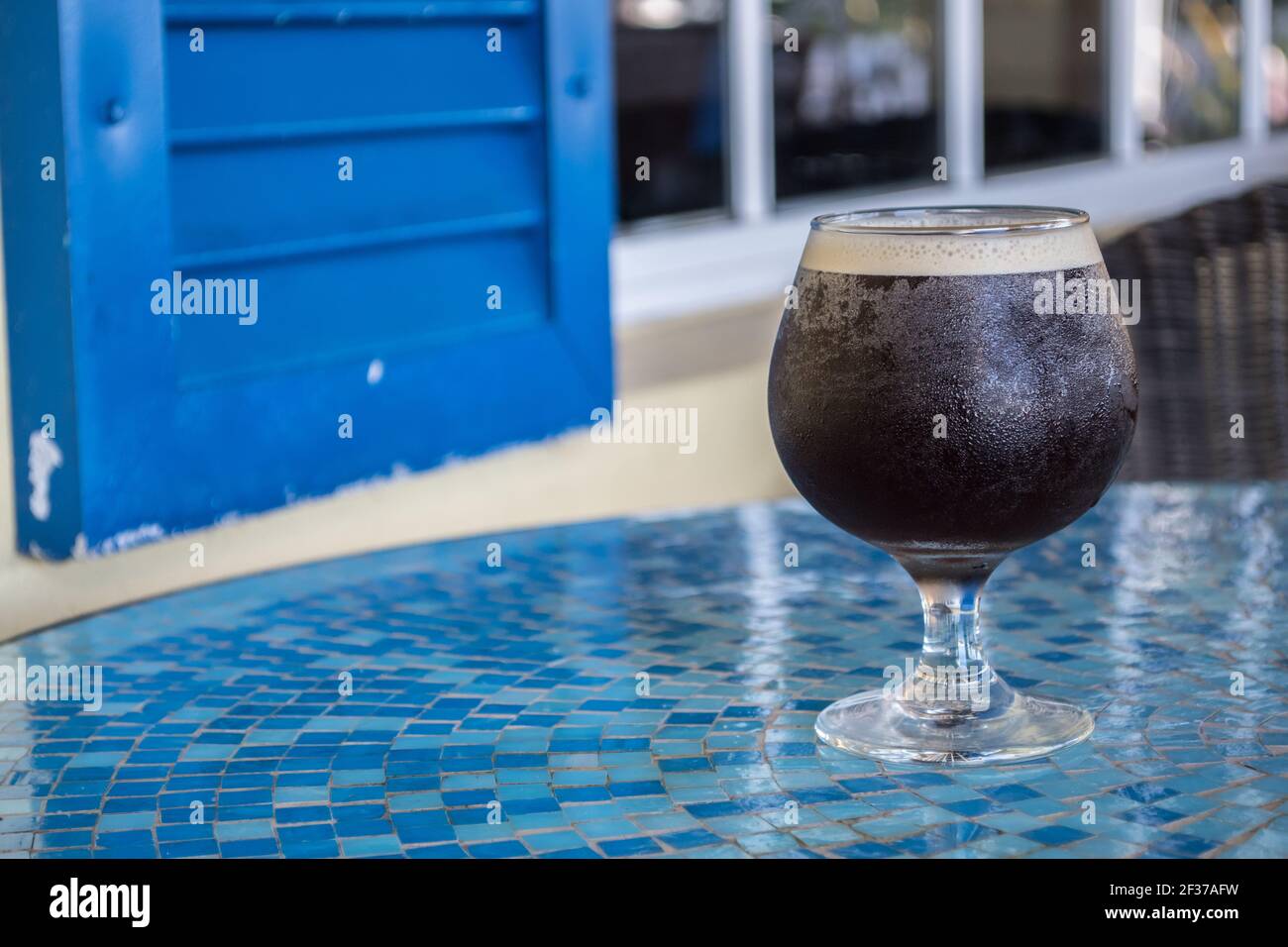 Bicchiere rotondo di caffè freddo con parte superiore in schiuma seduta su un tavolo di tegole a mosaico blu in un caffè all'aperto' Foto Stock