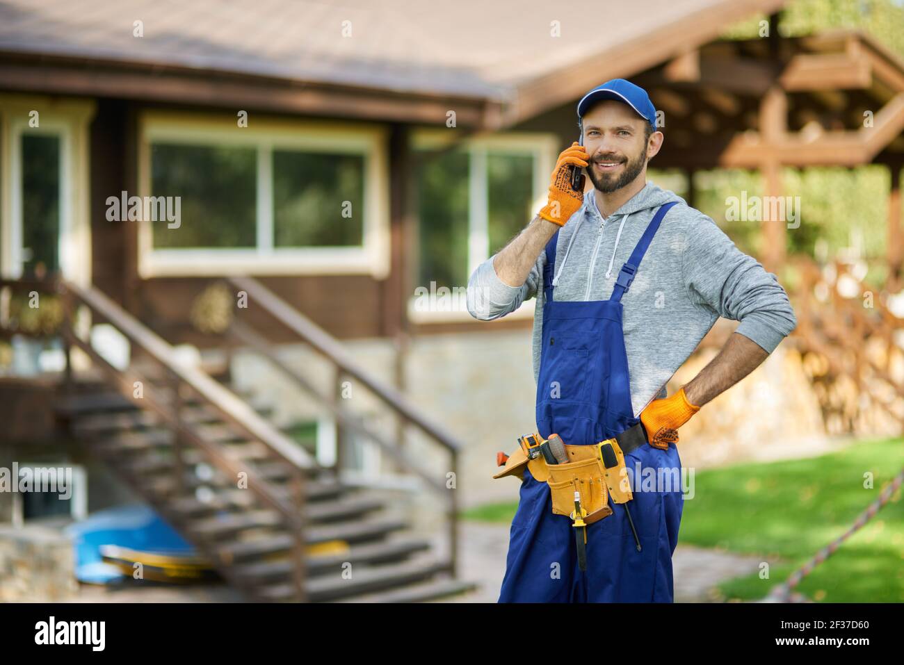 Allegro giovane costruttore maschile in uniforme sorridendo a macchina fotografica mentre si parla al telefono, in piedi all'aperto in casa cantiere su un caldo sole Foto Stock