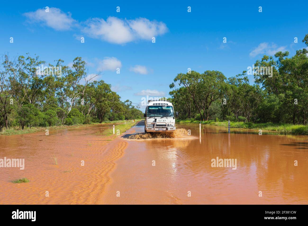 Toyota Coaster mini-bus guida attraverso inondazioni su una remota strada Outback coperto di fango rosso dopo una tempesta, vicino a Thallon, Queensland, QLD, Australia Foto Stock