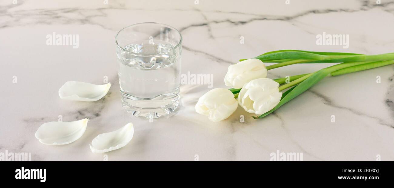 Un bicchiere di acqua potabile fresca purificata su un tavolo di marmo con fiori di tulipano bianchi. Copia spazio per il testo. Foto Stock