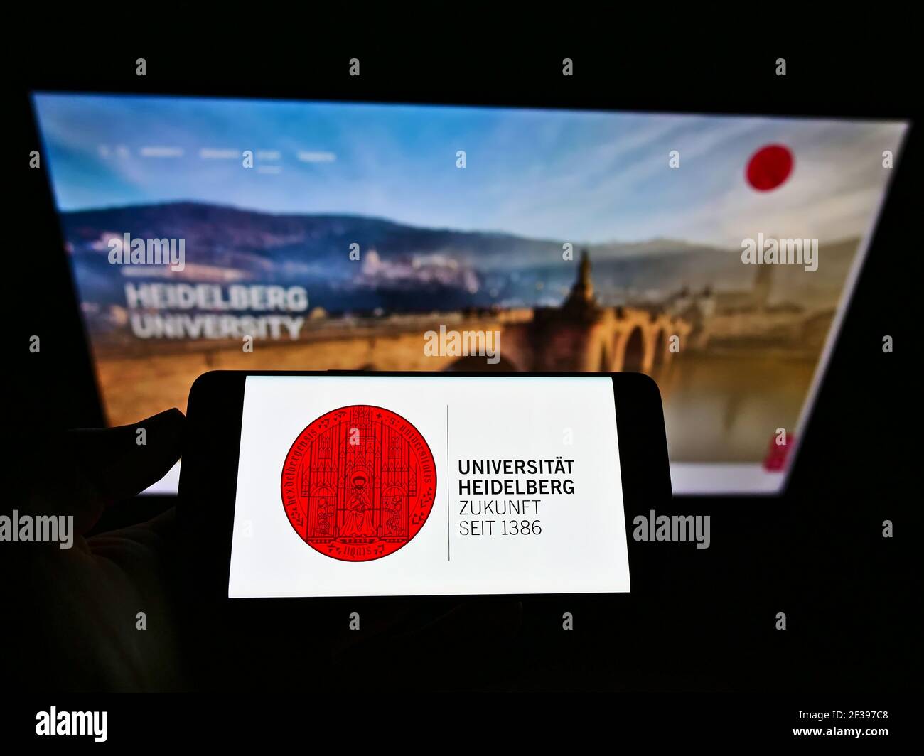 Persona che detiene il telefono cellulare con il logo dell'istituto tedesco di istruzione Heidelberg University sullo schermo di fronte alla pagina web. Mettere a fuoco il display del telefono. Foto Stock