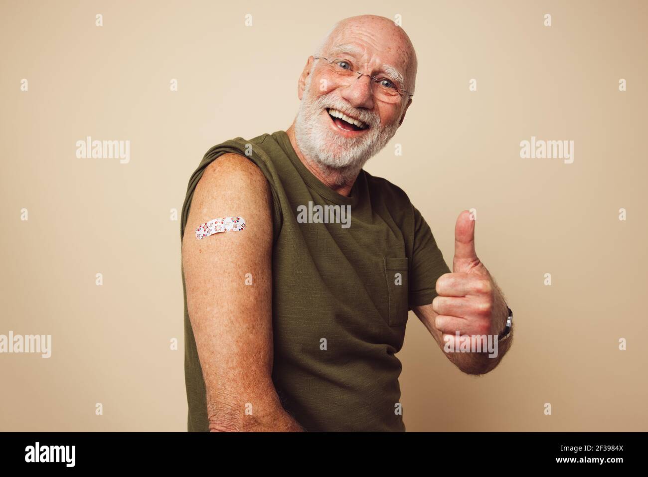 Ritratto di un maschio anziano sorridente e mostrante i pollici in su dopo avere ottenuto un vaccino. Uomo maturo con barba bianca seduta su fondo marrone sensazione Foto Stock