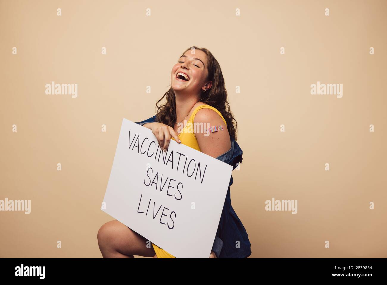Donna ridente con un banner che mostra 'la vaccinazione salva vitea'. Femmina dopo aver ottenuto la vaccinazione tenendo un banner su sfondo marrone. Foto Stock