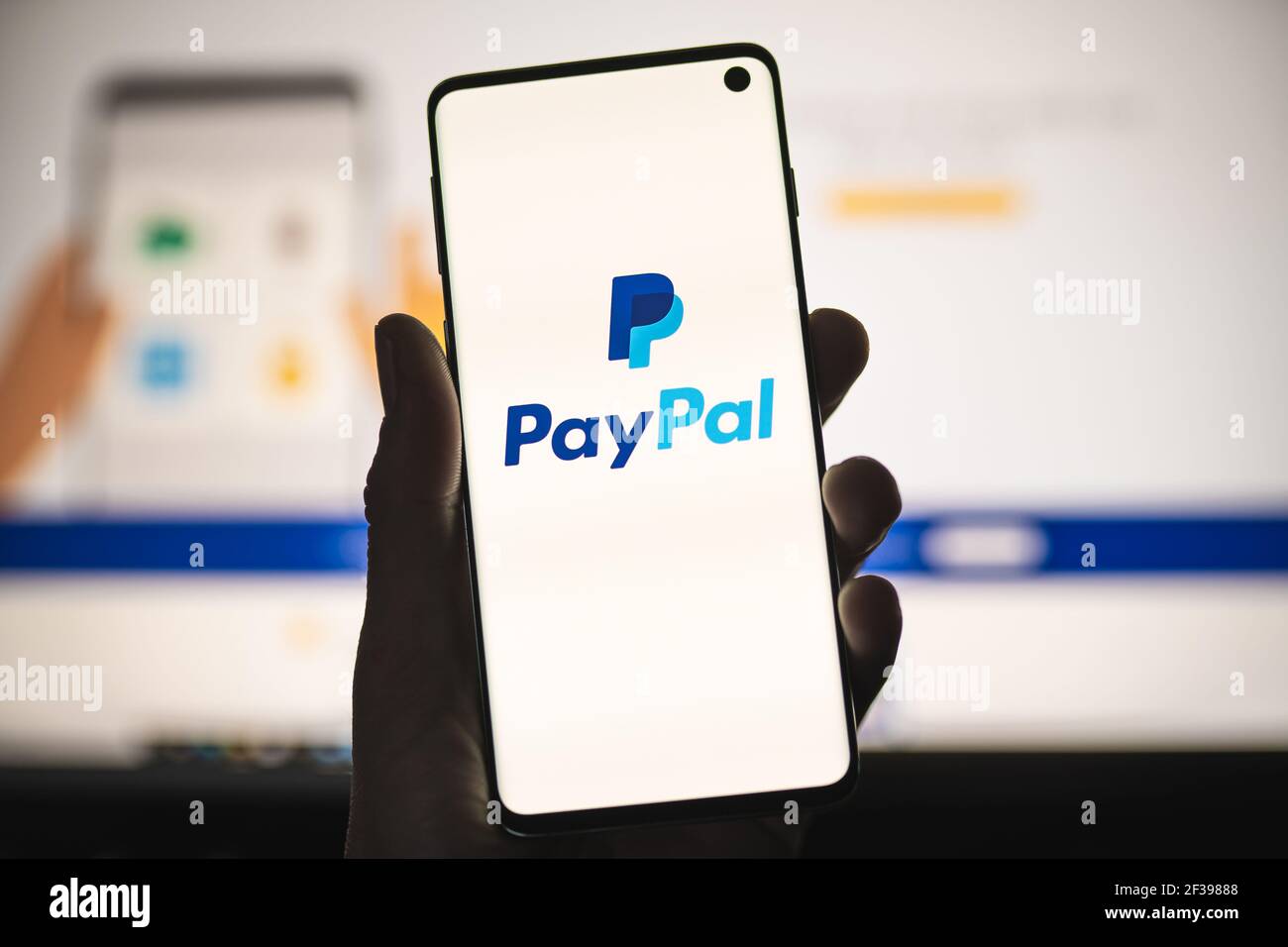 Uomo che tiene uno smartphone con il logo PayPal con sito web sfocato come sfondo. Sistema di pagamento online popolare, trasferimenti di denaro Foto Stock