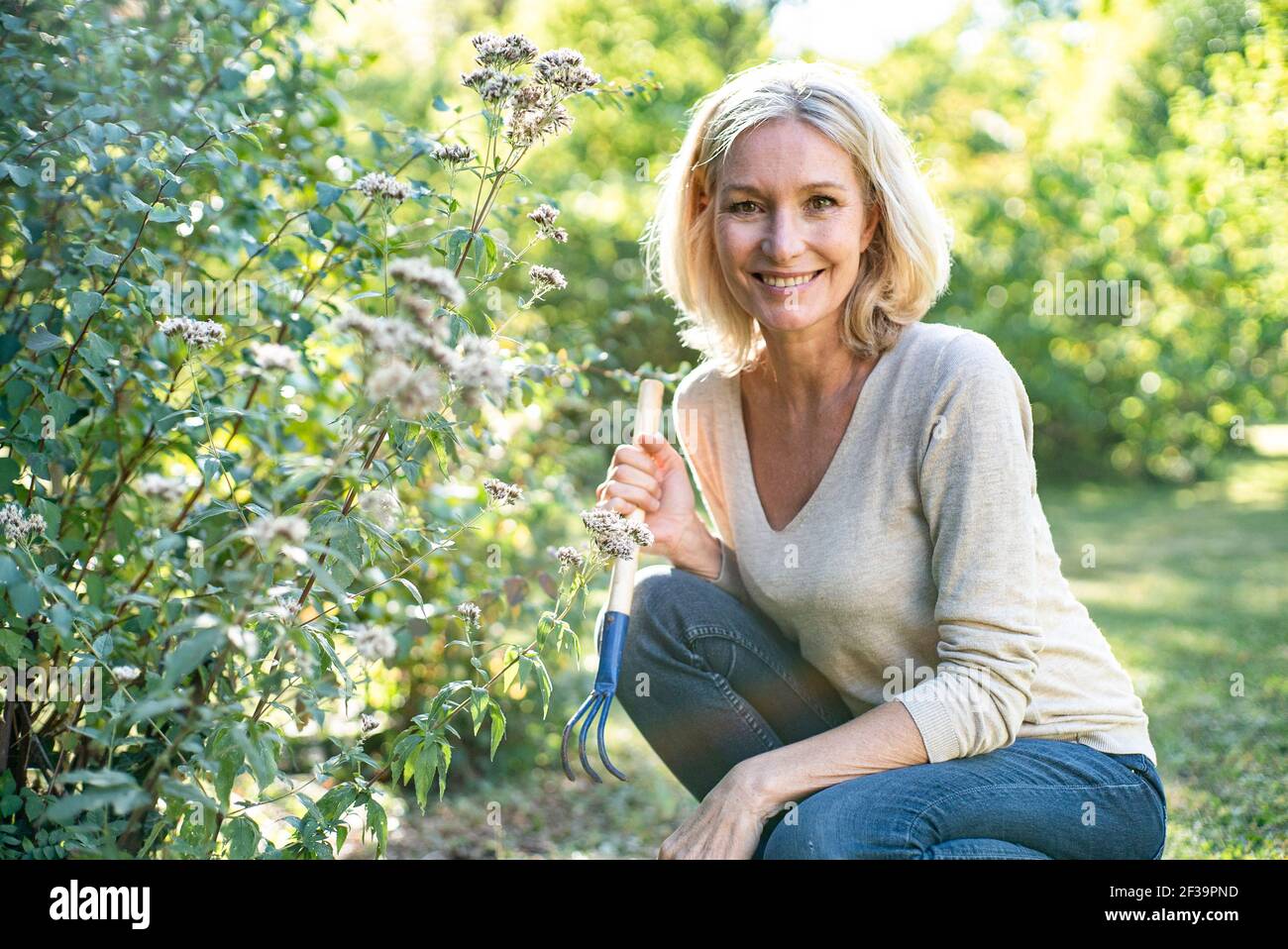Ritratto di donna matura sorridente con forchetta da giardino accovacciata in cortile Foto Stock