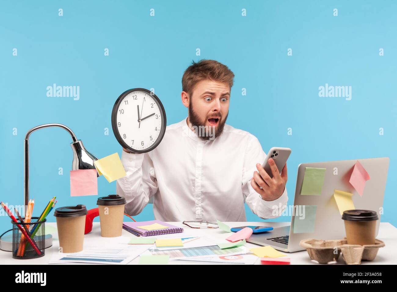 Terrorizzato uomo nervoso che lavora in ufficio tenendo orologio da parete e guardando lo smartphone display con paura, in ritardo su appuntamento, mancante riunione di affari. IND Foto Stock