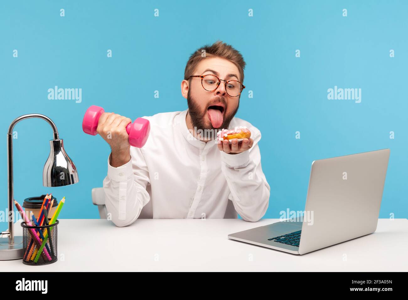 Lavoratore di ufficio di uomo bearded divertente che vuole leccarsi ciambella tenendo in mano il dumbbell seduto sul posto di lavoro, scelta difficile tra sport e mangiare malsano, io Foto Stock