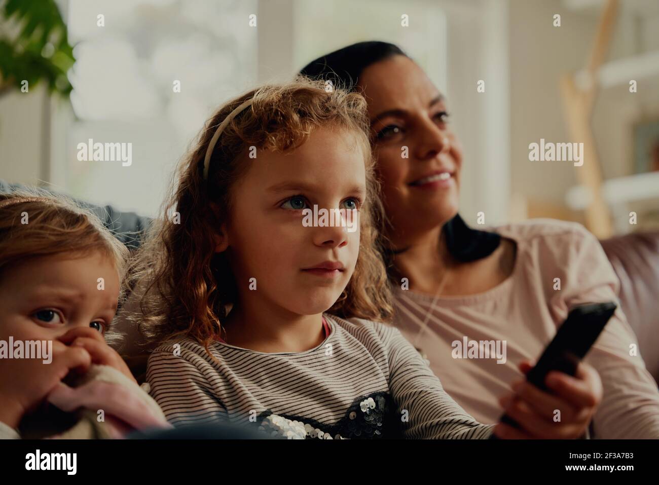Ragazza concentrata che tiene il telecomando guardando la televisione con la madre e. sorella a casa Foto Stock