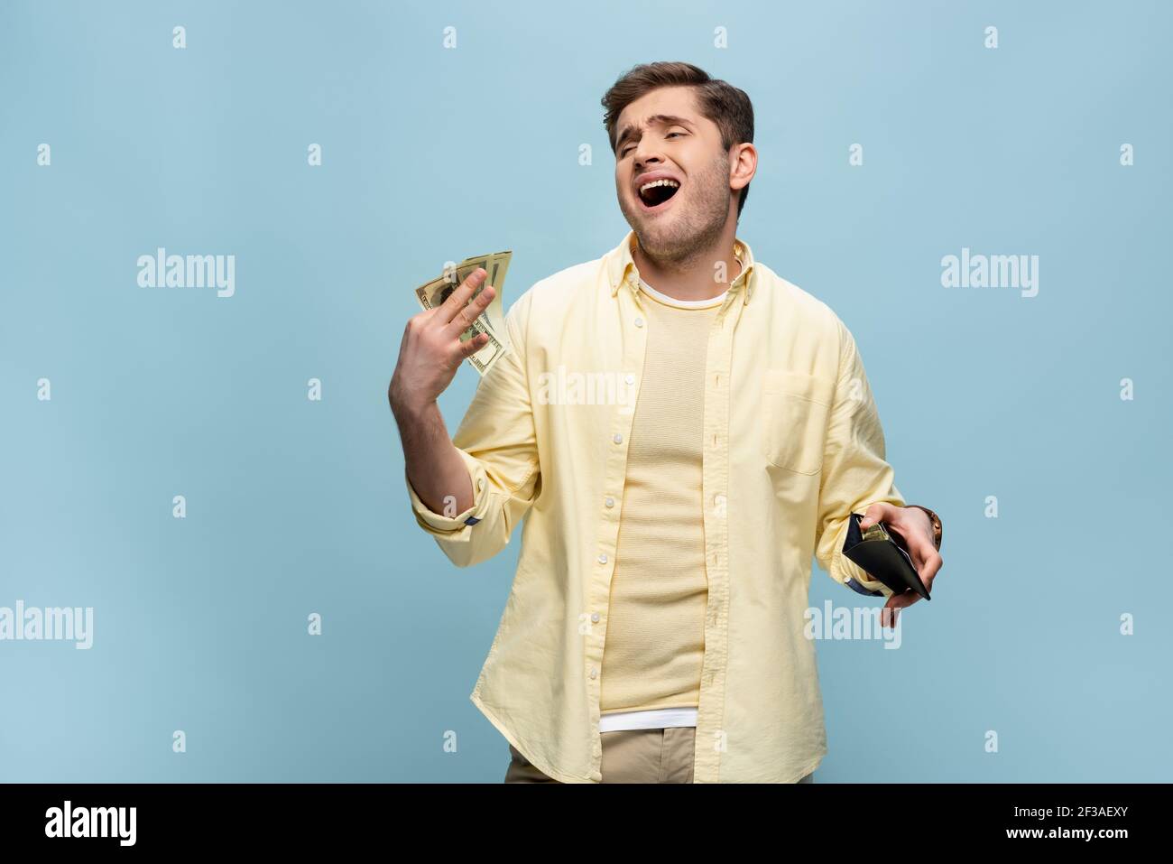 giovane ricco in camicia gialla che tiene il portafoglio e sventolando dollari isolati in blu Foto Stock