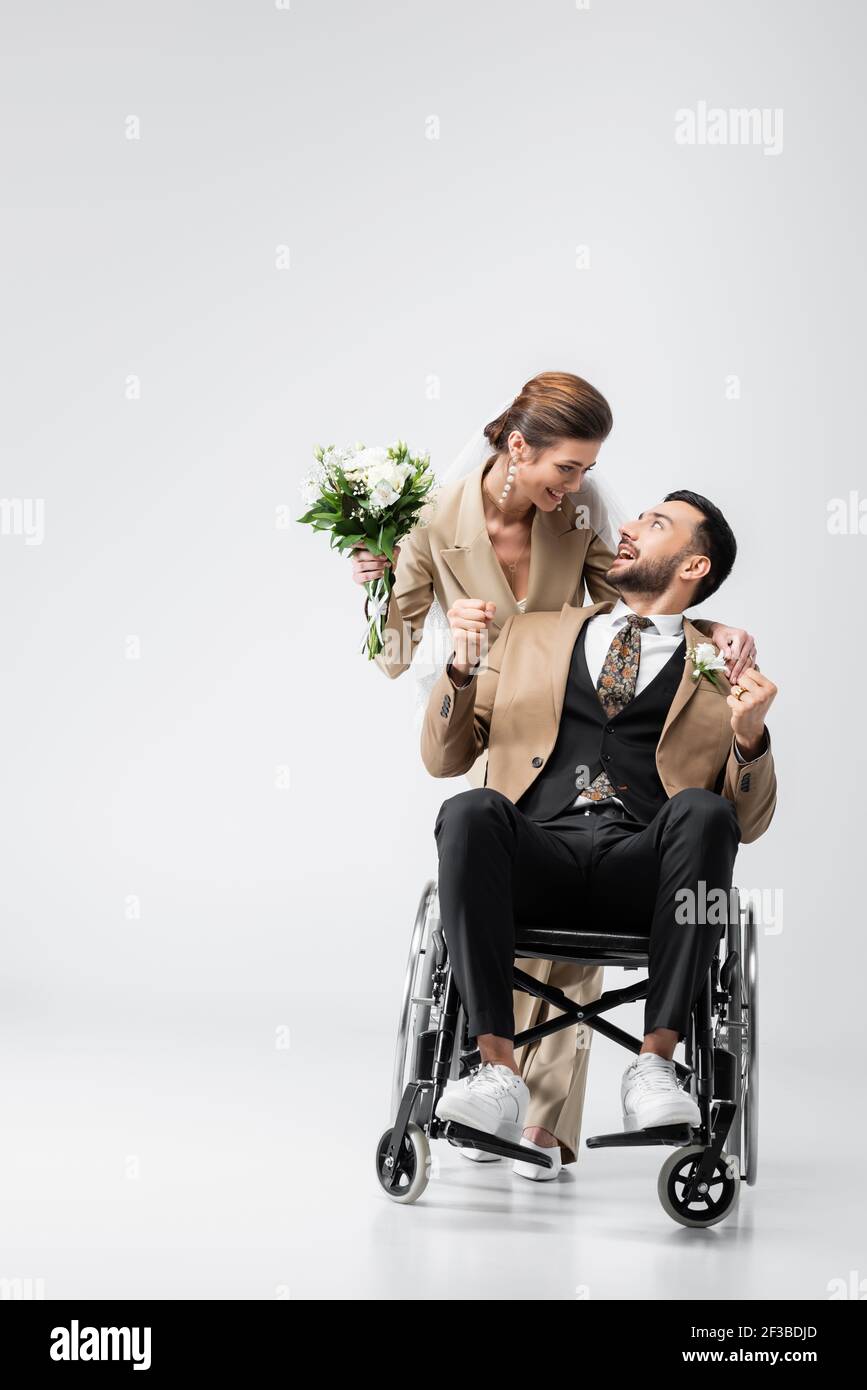 fiance musulmana handicappato che mostra la vittoria gesto vicino sposa felice con bouquet di nozze su grigio Foto Stock