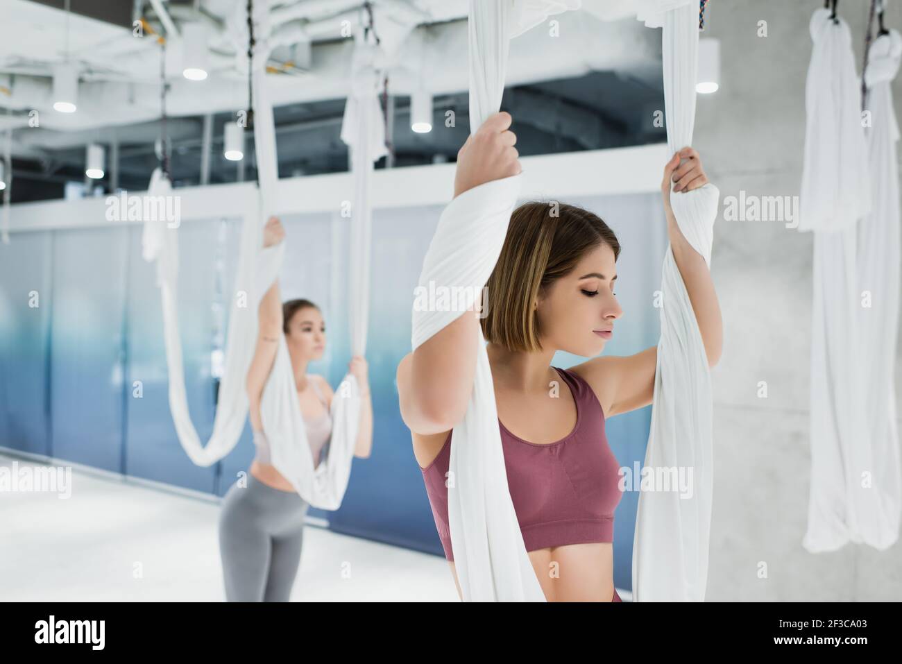 giovani donne sportive che allungano le braccia con cinghie di yoga mosca in centro fitness, sfondo sfocato Foto Stock
