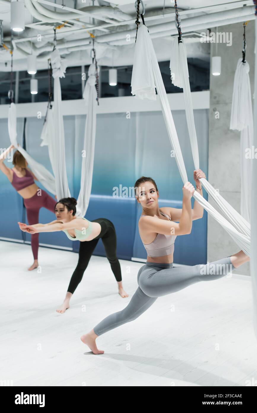 Donna sportiva a piedi nudi che lavora con amaca yoga mosca vicino alle donne su sfondo sfocato nel centro fitness Foto Stock