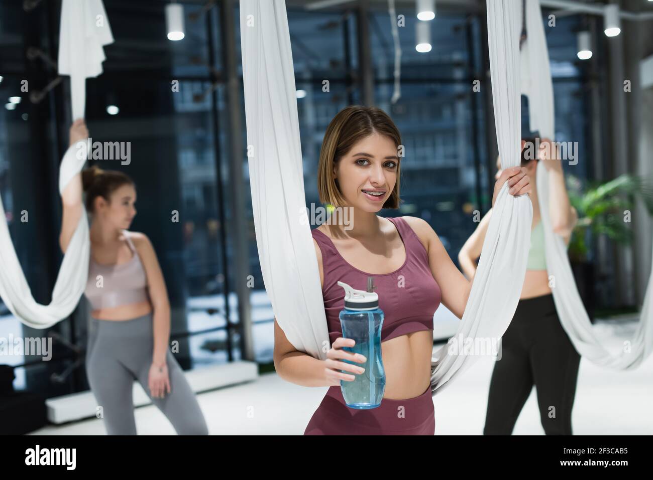 Donna allegra che sorride mentre tiene una bottiglia di sport e aero yoga cinturini nel centro fitness Foto Stock
