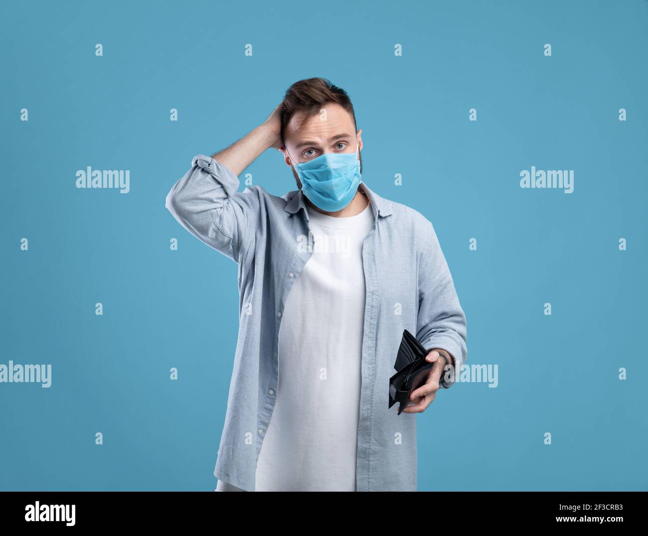 Fallimento durante il covid. Uomo millenario in maschera di faccia che tiene il portafoglio vuoto e graffiando la testa su sfondo blu Foto Stock