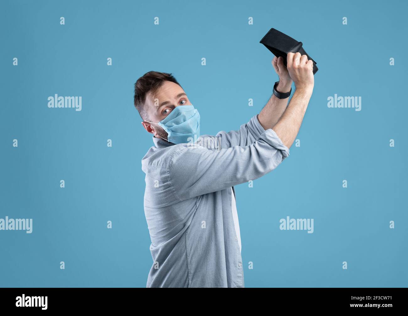 Disperato uomo caucasico in maschera protettiva scuotendo portafoglio vuoto, non avendo soldi durante covid su sfondo blu Foto Stock