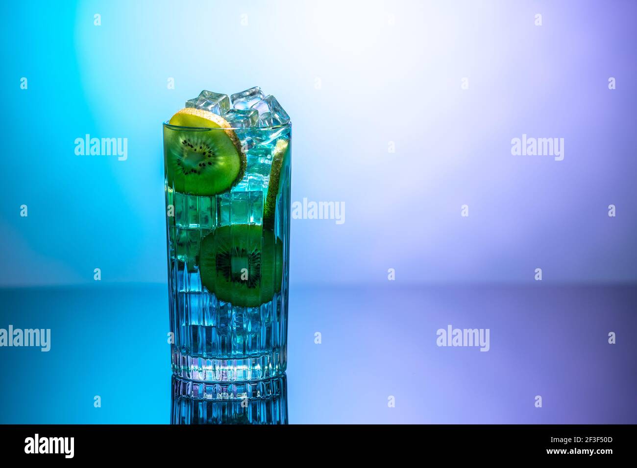 Un bicchiere d'acqua con ghiaccio e fette di kiwi in piedi su un tavolo a specchio in luce al neon. Concetto di disintossicazione o disintossicazione del corpo Foto Stock