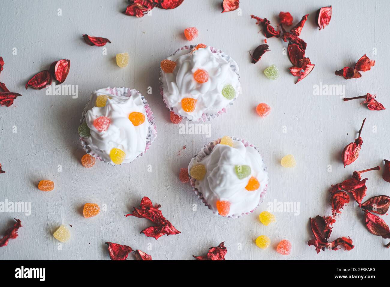 Tre cupcake con fagioli di gelatina su sfondo bianco. Pezzi di rose asciutte intorno a loro. Focalizzazione selettiva su aree diverse. Foto Stock