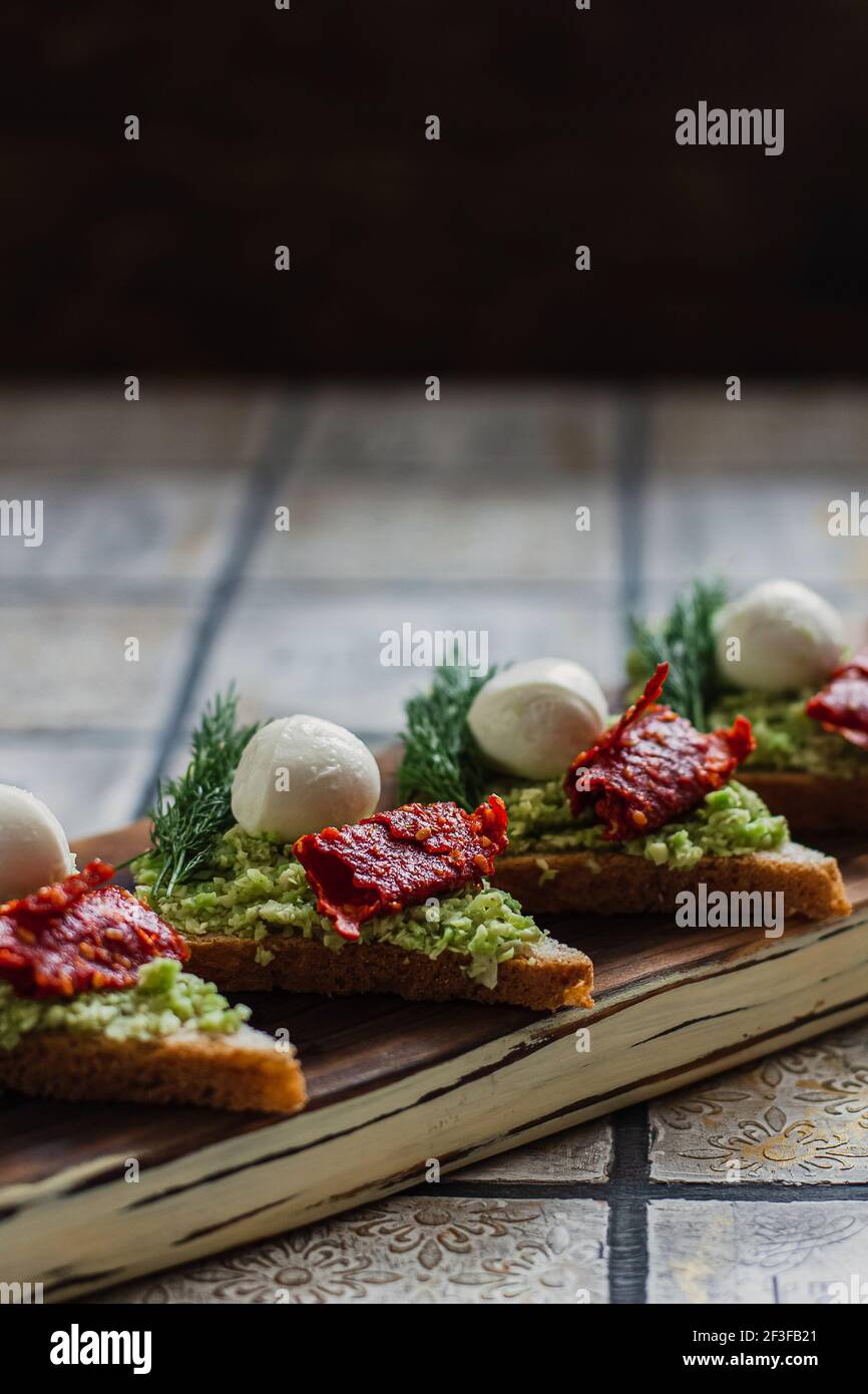 Mini crostini triangolari bruschetta con avocado, mozzarella e tomatos essiccati sulla tavola rustica sullo sfondo piastrellato d'annata Foto Stock
