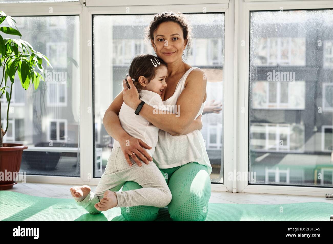 Affascinante giovane donna, madre, abbracciando la sua figlia carina mentre si siede su un tappeto di yoga sullo sfondo di grandi finestre. Esercitarsi a casa su un b Foto Stock