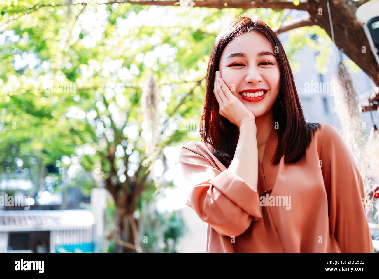 Ritratto di giovane asiatica bella donna in piedi in città con un sorriso togoloso con le mani sulla guancia guardando la macchina fotografica Foto Stock