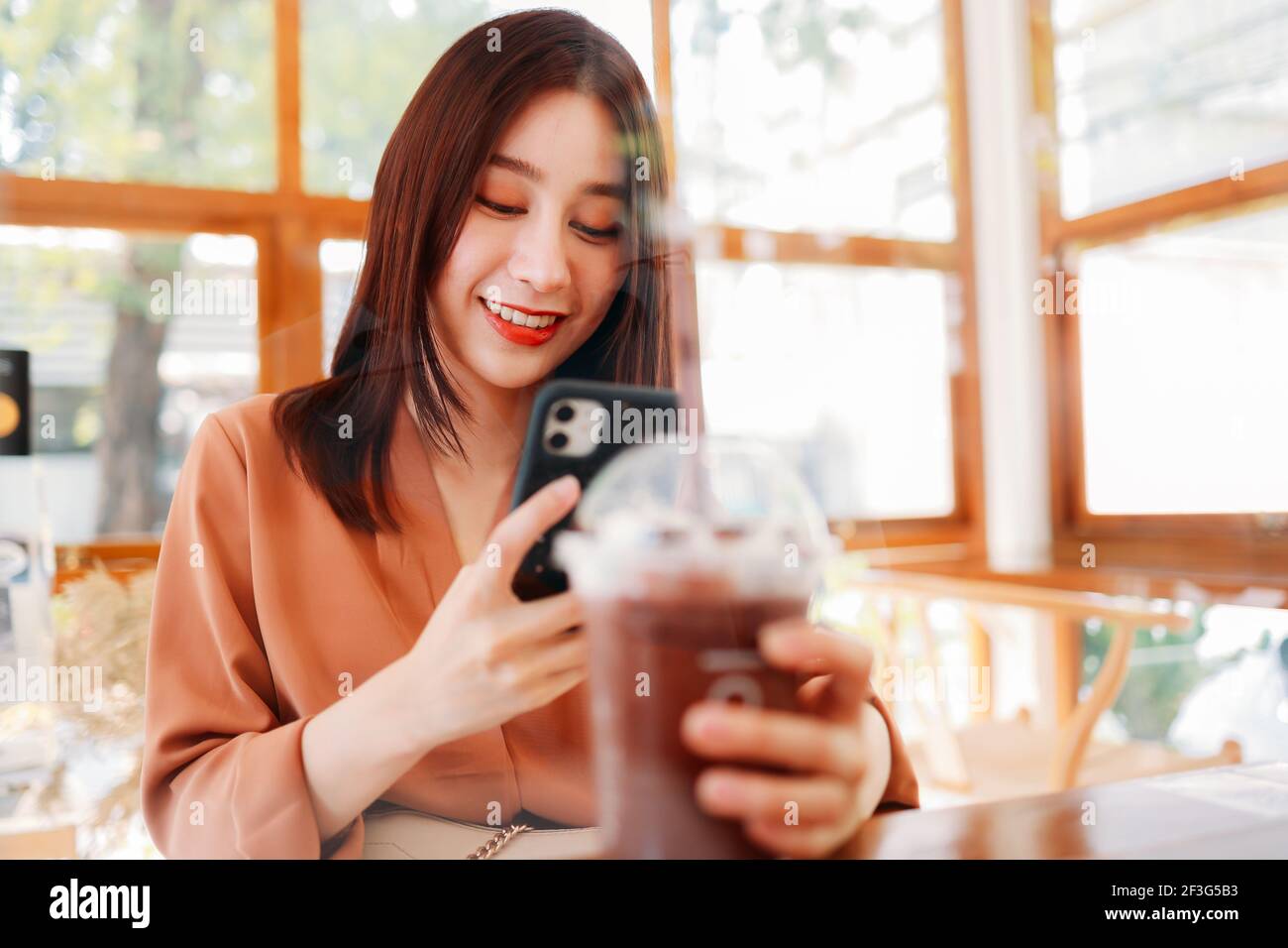 Sorridente bella giovane asiatica blogger cibo femminile tenendo una presa allontanate il bicchiere di frullato e frullato mentre scattate una foto telefono Foto Stock