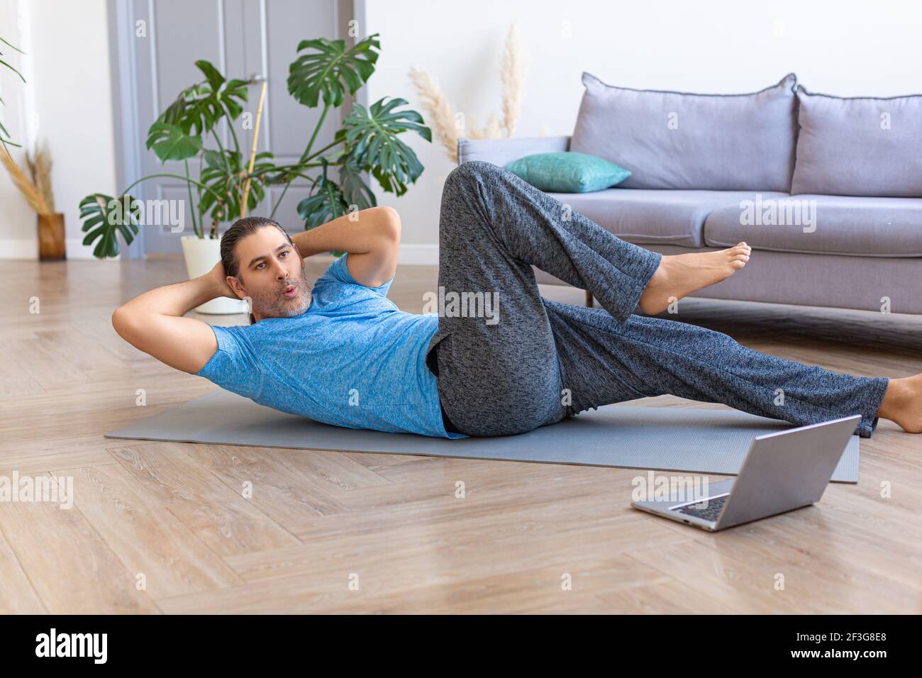 Uomo di mezza età che pratica gli scricci dell'abs, che addestrano i muscoli addominali sul pavimento mentre guarda il video di idoneità in linea sul laptop. Allenamento domestico e healt Foto Stock