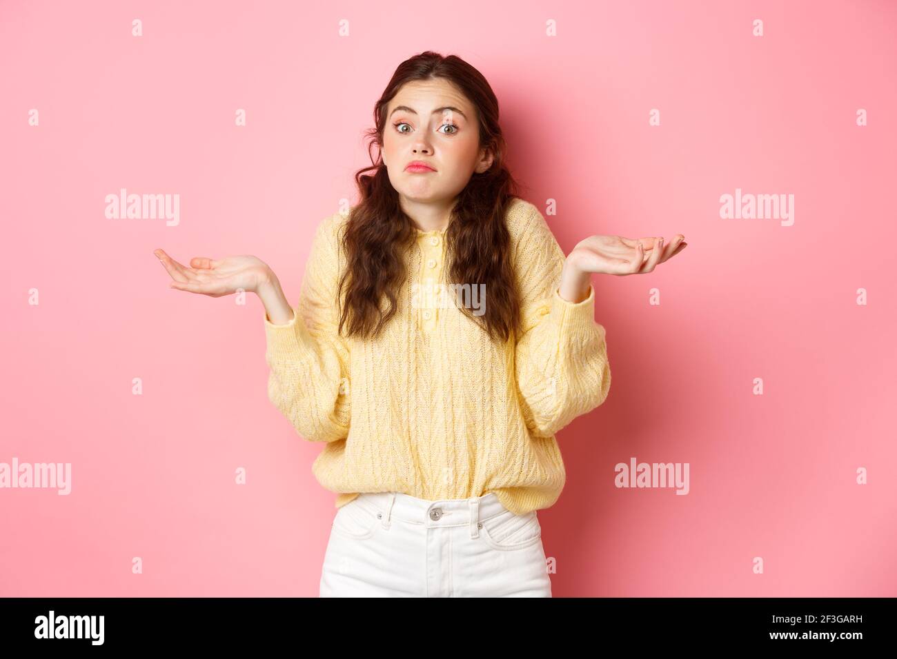 Non lo so. Ragazza indecisa che si infrange le spalle e che guarda ignaro alla macchina fotografica, in piedi indecisa contro sfondo rosa Foto Stock