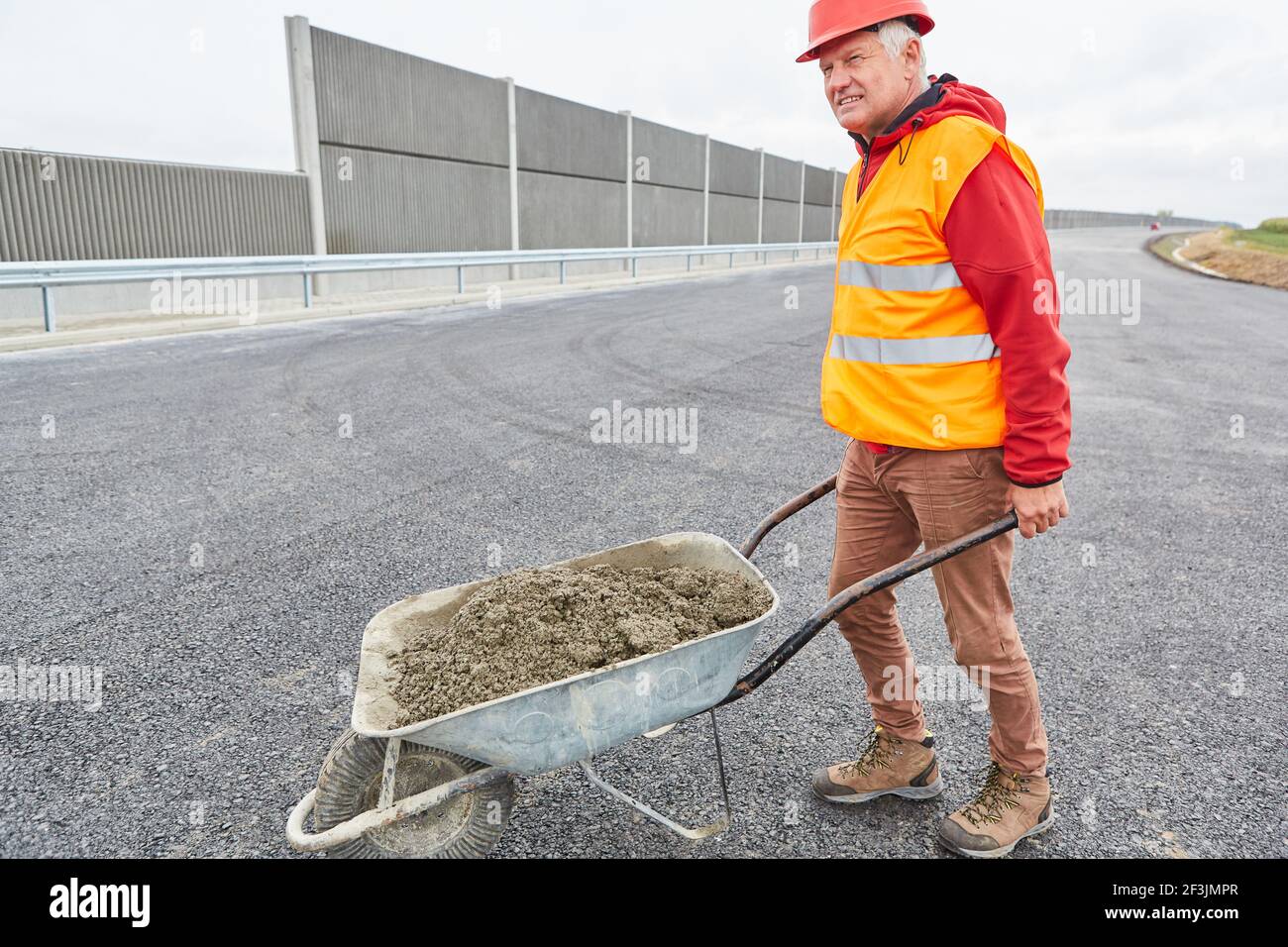 Operaio di costruzioni con carriola piena di calcestruzzo sul cantiere durante l'espansione della tangenziale Foto Stock