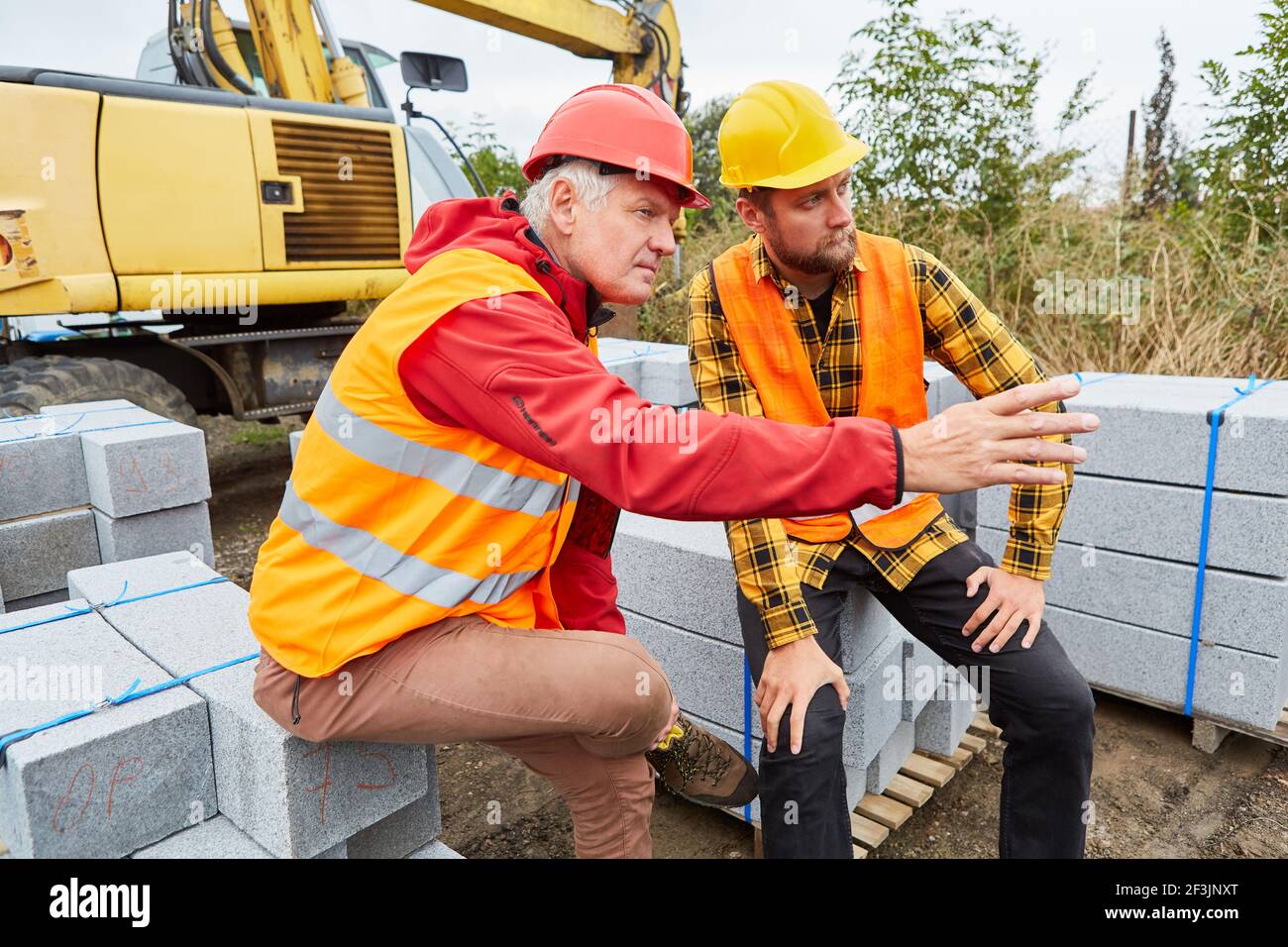 Due operai edili sul cantiere della casa con materiali da costruzione e escavatore Foto Stock